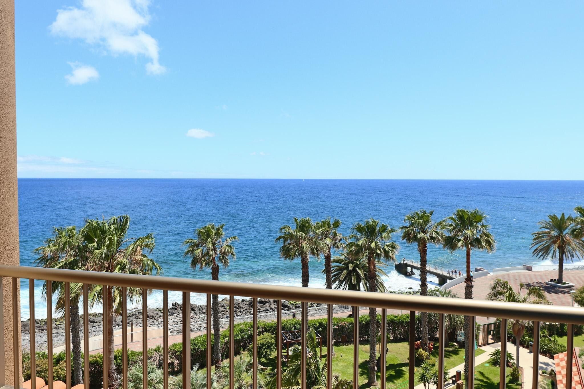 Un balcón con vista al océano, palmeras y una playa rocosa.