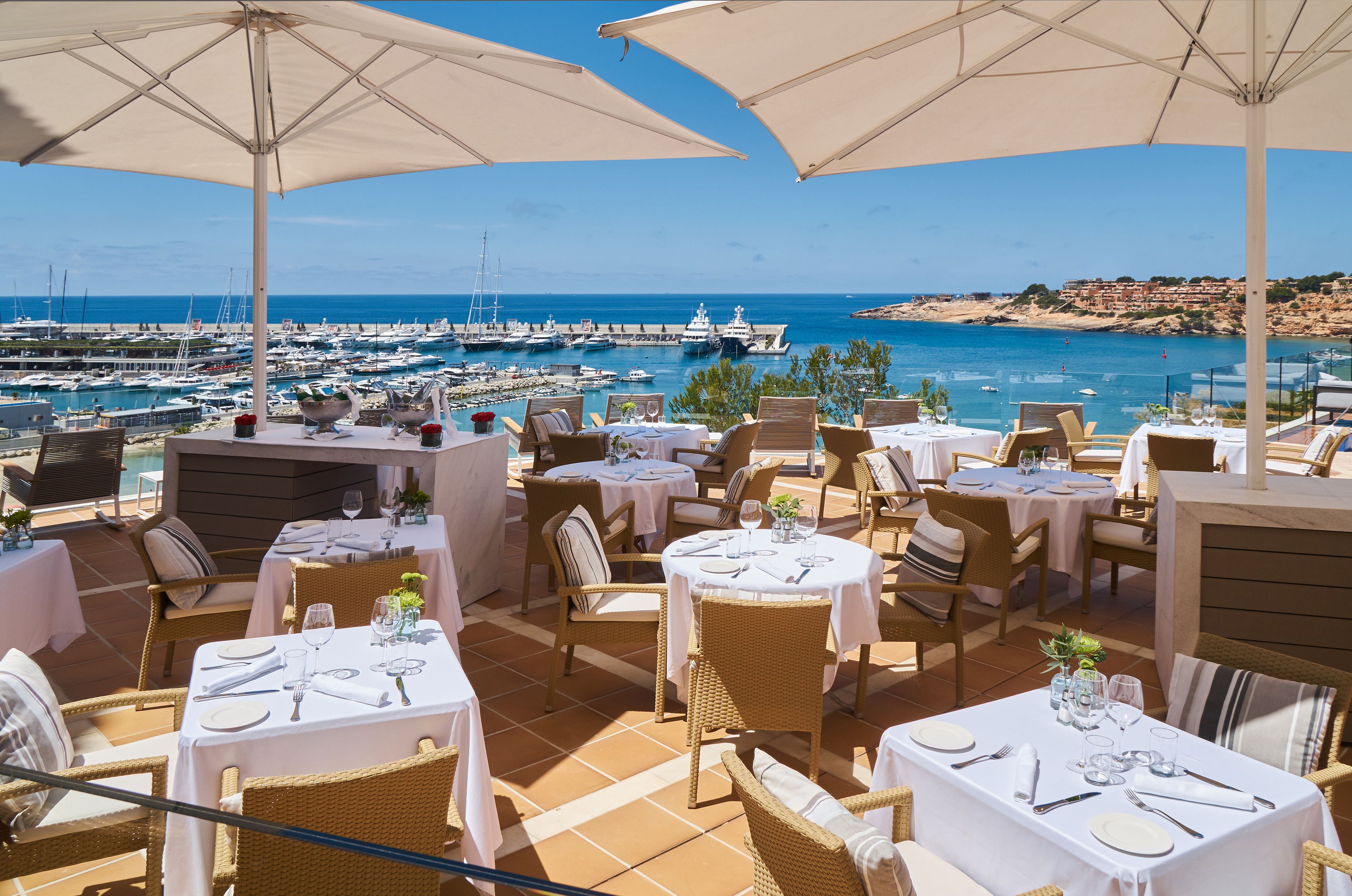 An outdoor dining area with tables set for two, overlooking a marina and the sea.