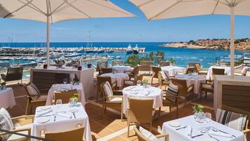 An outdoor dining area with tables set for two, overlooking a marina and the sea.