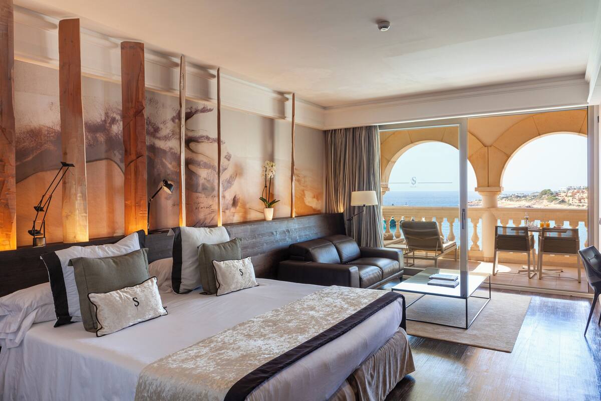 A hotel room with a large bed, a sofa, a desk, and a balcony with a view of the sea.