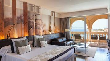 A hotel room with a large bed, a sofa, a desk, and a balcony with a view of the sea.