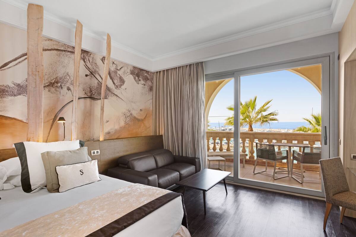 A hotel room with a bed, a sofa, a table, and a balcony with a view of the beach.