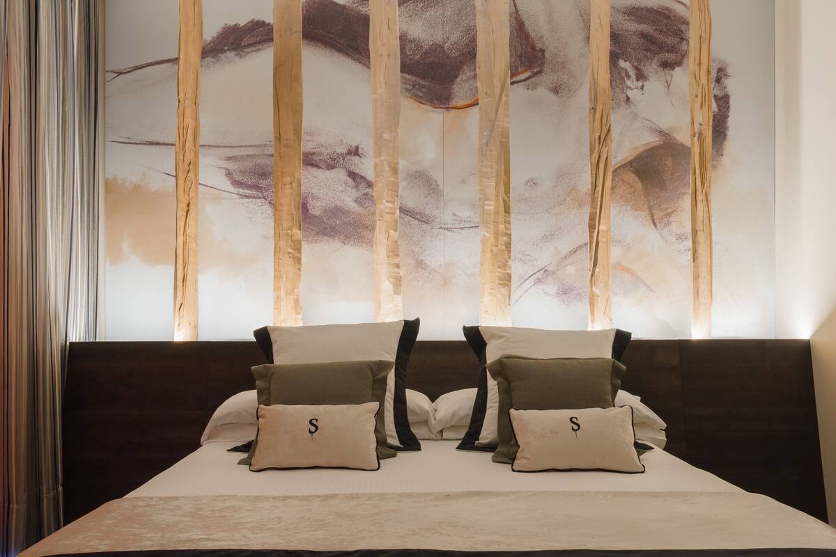 A bedroom with a large bed, two pillows with a letter 'S', and a wall with a large abstract painting.