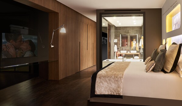 Un dormitorio moderno con una cama grande, un televisor y un baño visible a través de una puerta abierta.