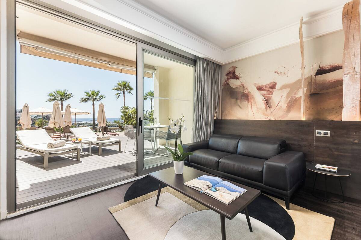 A modern living room with a large sliding glass door leading to a balcony with lounge chairs and umbrellas, a black leather sofa, and a coffee table with a magazine.