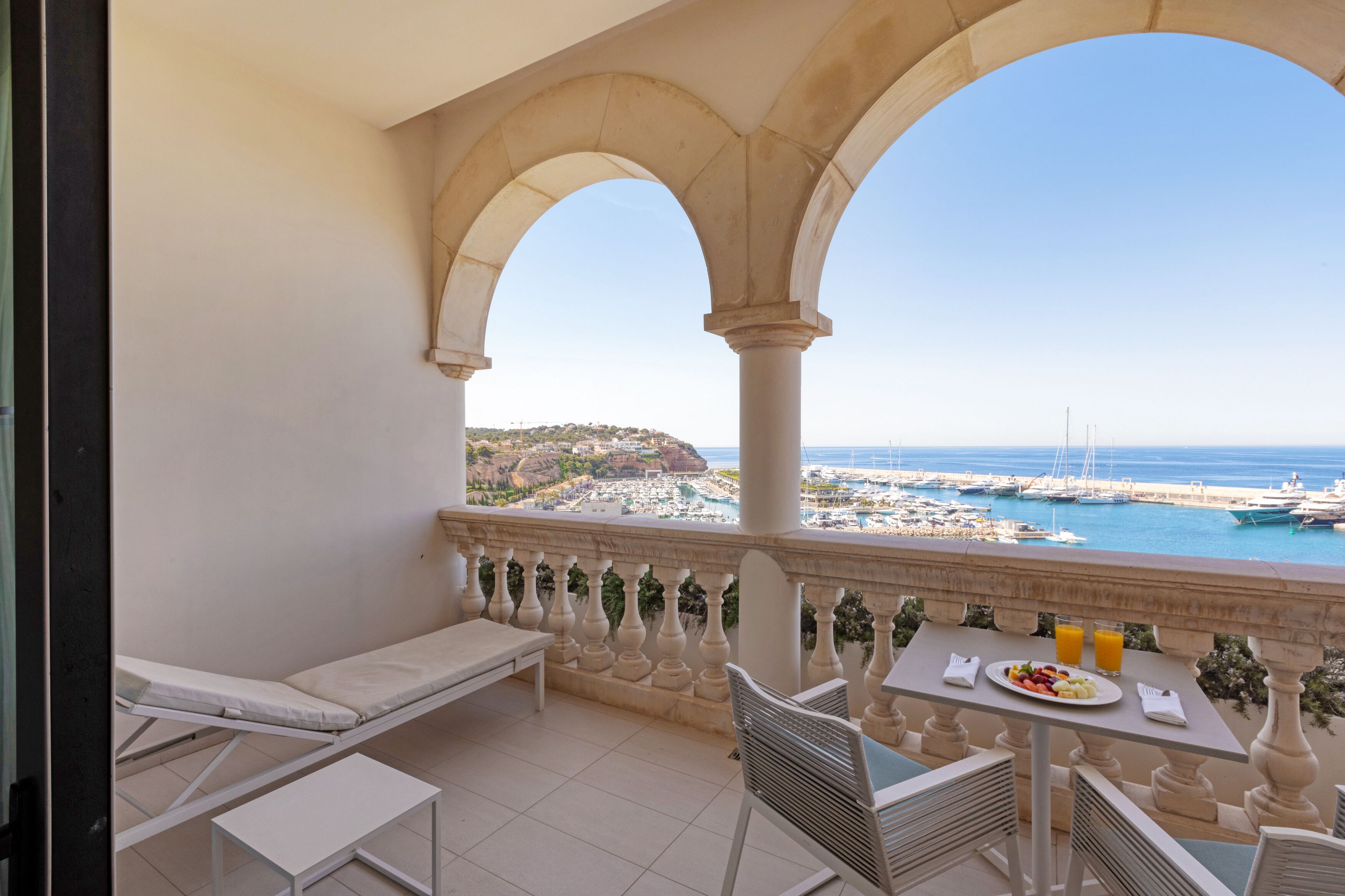 Ein Balkon mit Meerblick, ein Esstisch mit Speisen und Getränken sowie eine Liege.
