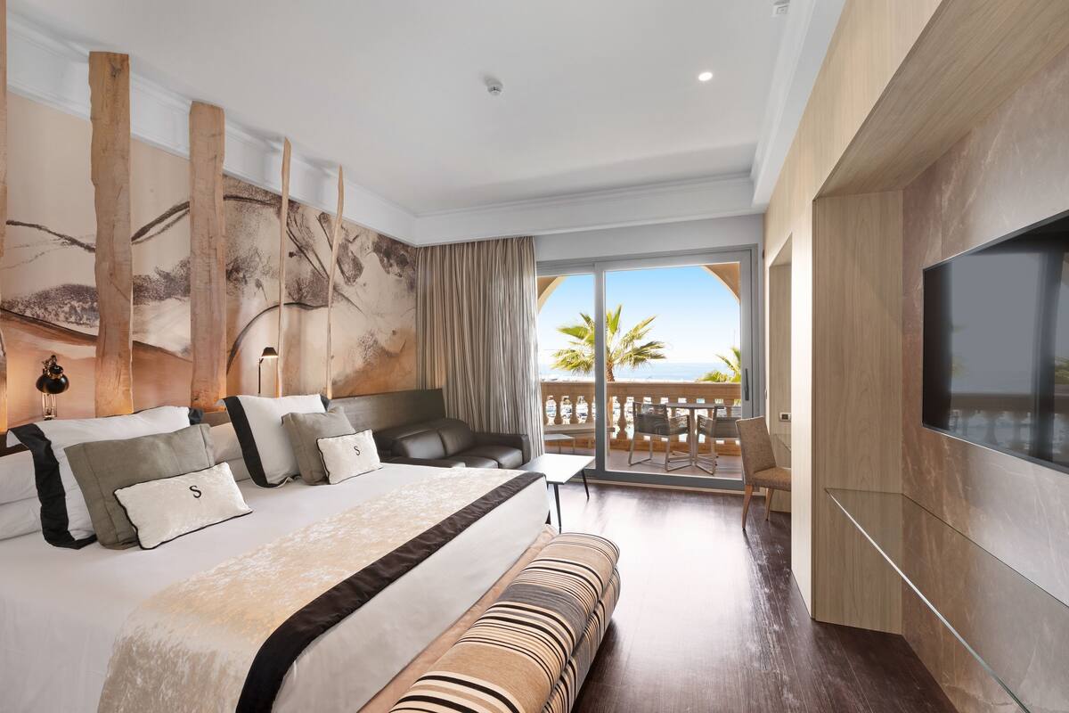 A modern hotel room with a large bed, a TV, a balcony with a view, and a wooden headboard.