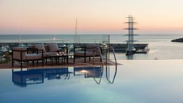 Una piscina infinita con sillones de descanso y una mesa, con vistas a un puerto deportivo con barcos y un barco.