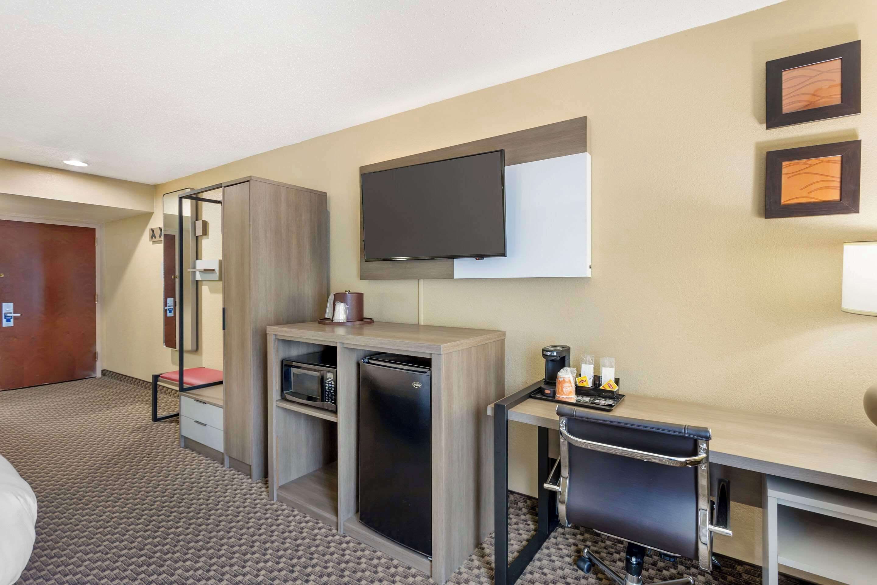 A hotel room with a bed, a desk with a chair, a refrigerator, a microwave, a flat-screen TV mounted on the wall, and a small coffee station.