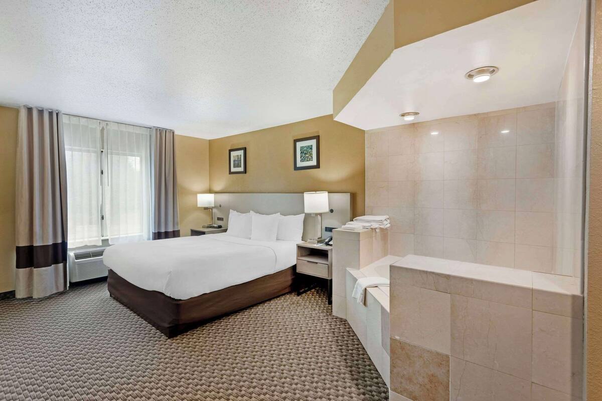 A hotel room with a large bed, a bedside table with a lamp, a bathroom with a bathtub, and a window with curtains.