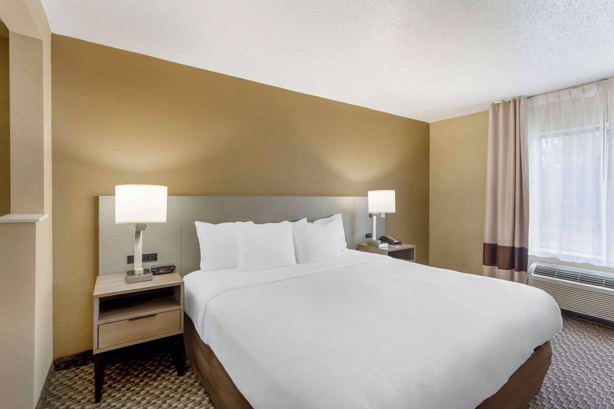 A hotel room with a large bed, two bedside tables with lamps, a nightstand, and a window with curtains.