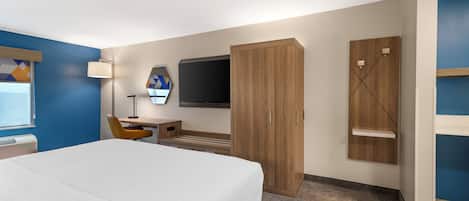 A hotel room with a bed, desk, chair, TV, wardrobe, and a small shelf.