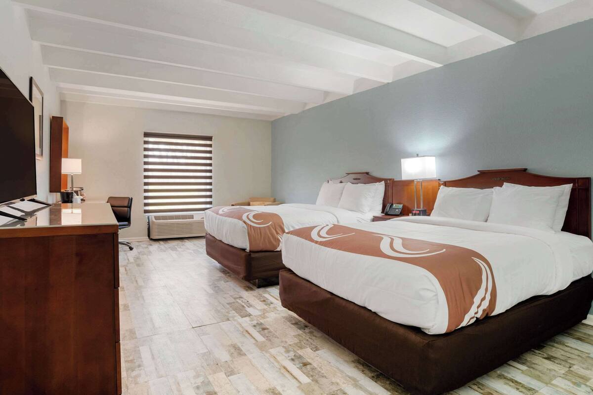 Quarto de hotel com uma cama grande, uma escrivaninha e uma cadeira. Há uma janela com persianas, uma luminária e um telefone na escrivaninha.