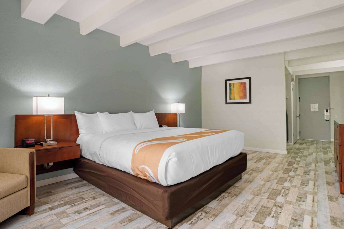 Quarto de hotel com uma cama grande, duas mesas de cabeceira com luminárias, uma cadeira e uma porta que dá para outro quarto.