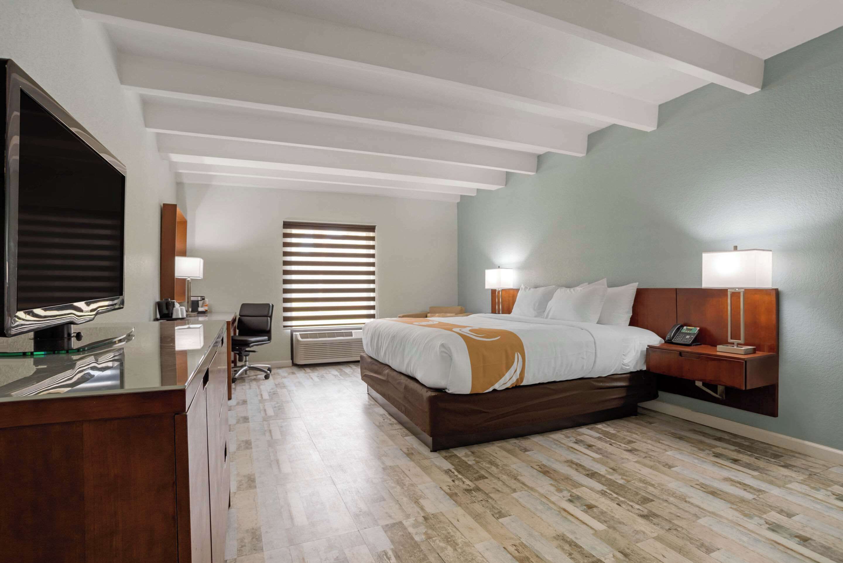 A hotel room with a bed, bedside table, lamp, TV, and desk.