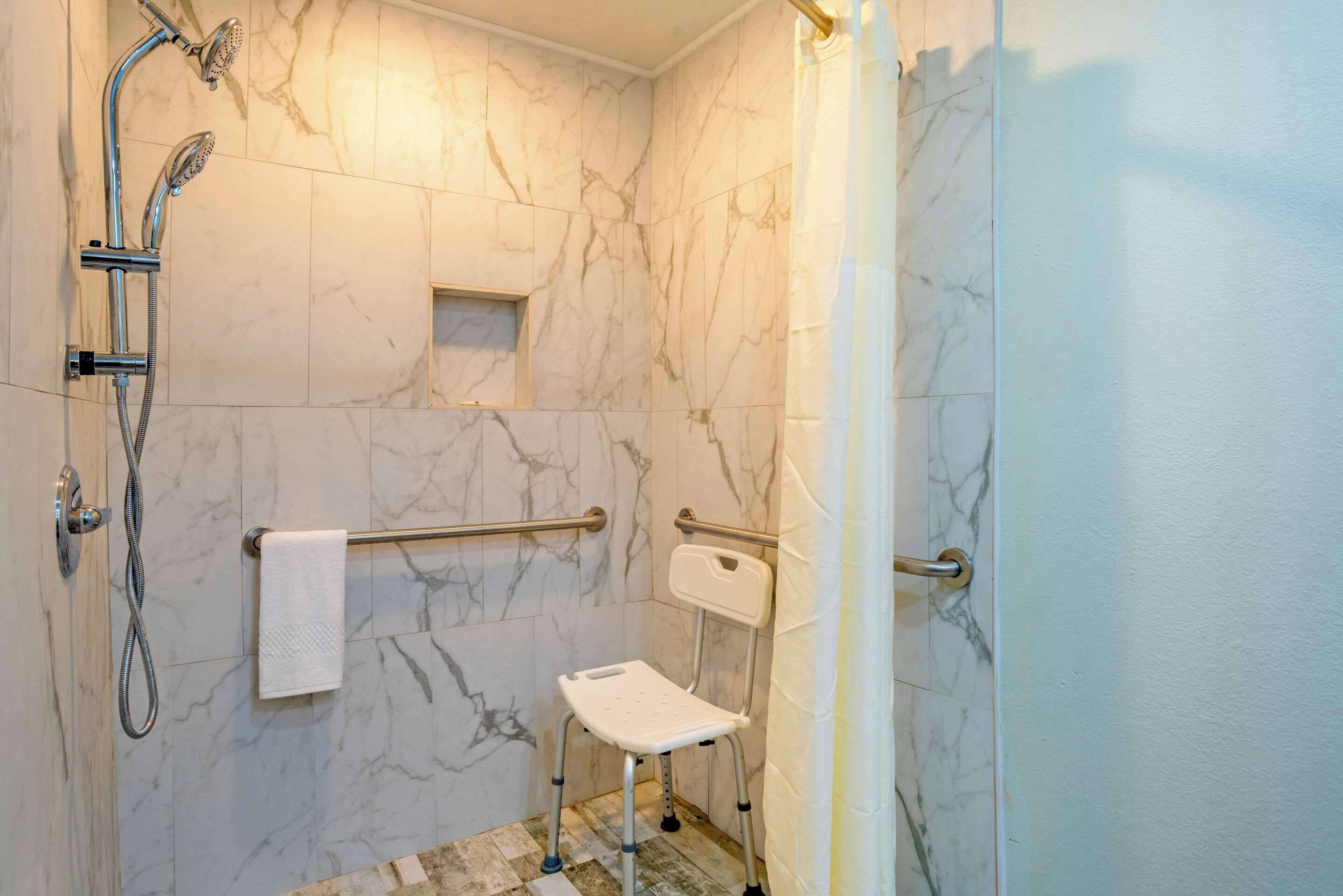 A bathroom with marble walls, a shower with a handheld showerhead, a grab bar, and a small step stool.