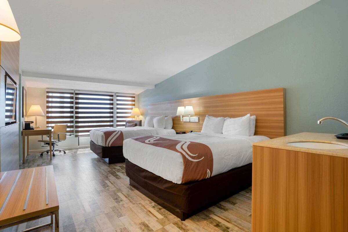 Quarto de hotel com duas camas, uma escrivaninha, uma cadeira, uma luminária de mesa, uma mesa de cabeceira e um banheiro.