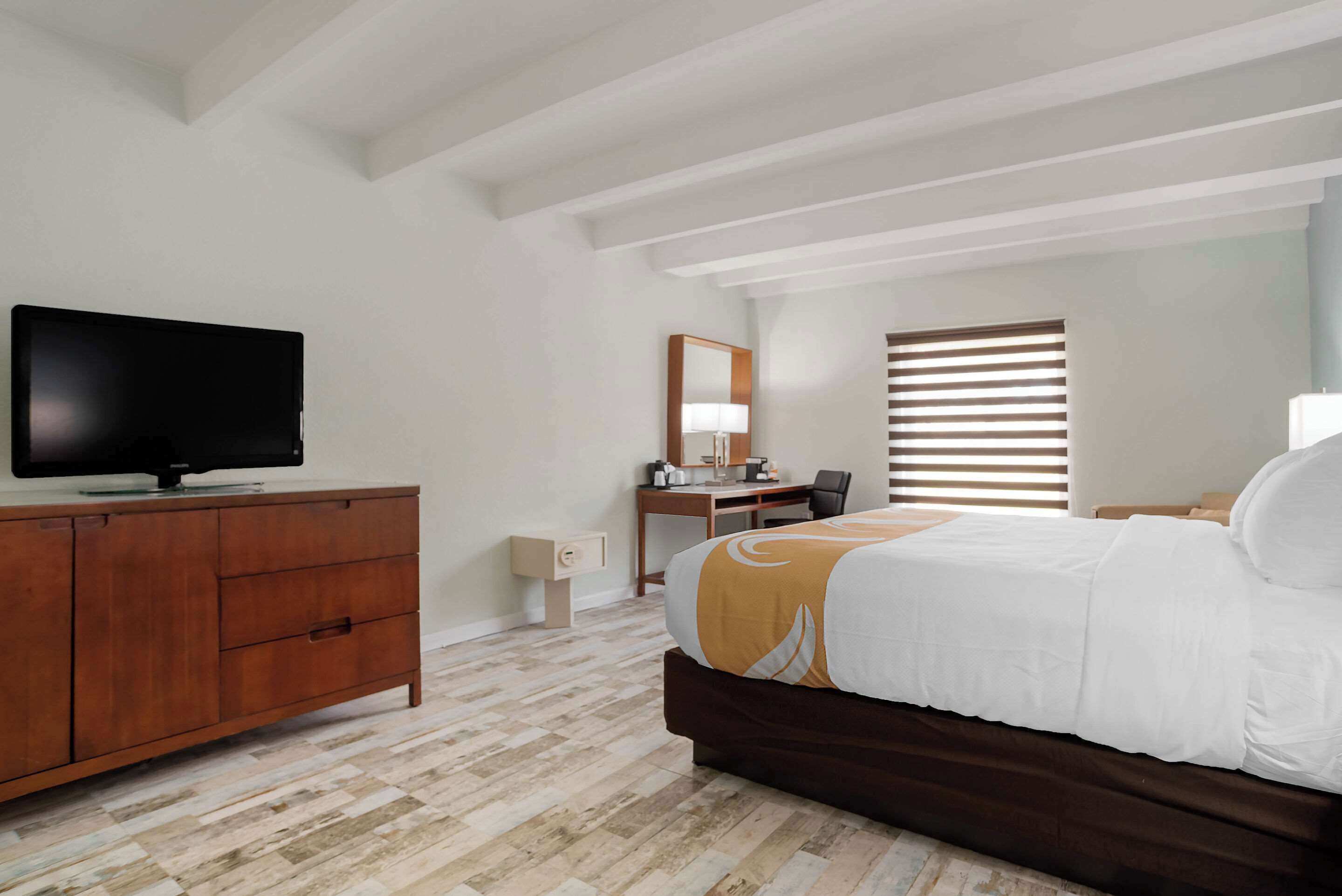 A hotel room with a bed, a TV on a wooden dresser, a desk with a chair, and a window with blinds.
