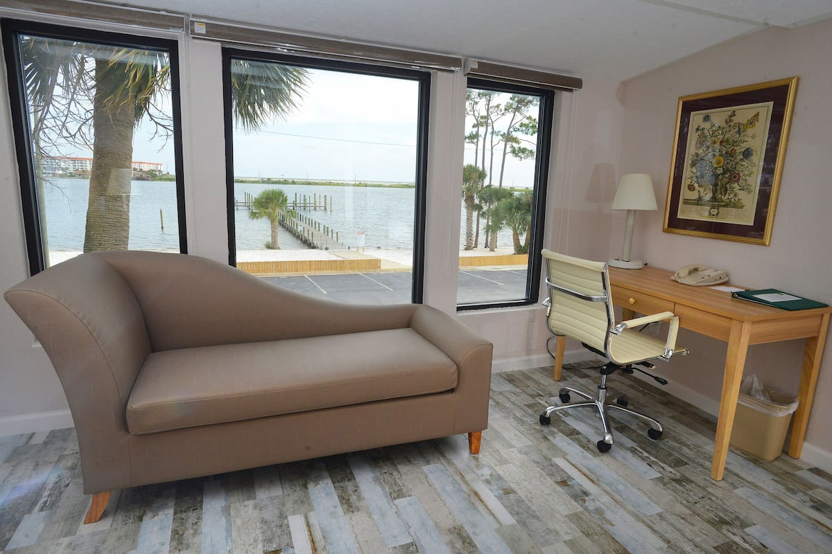 Quarto com sofá bege, escrivaninha de madeira e uma cadeira. Janelas amplas oferecem vista para a água e palmeiras.