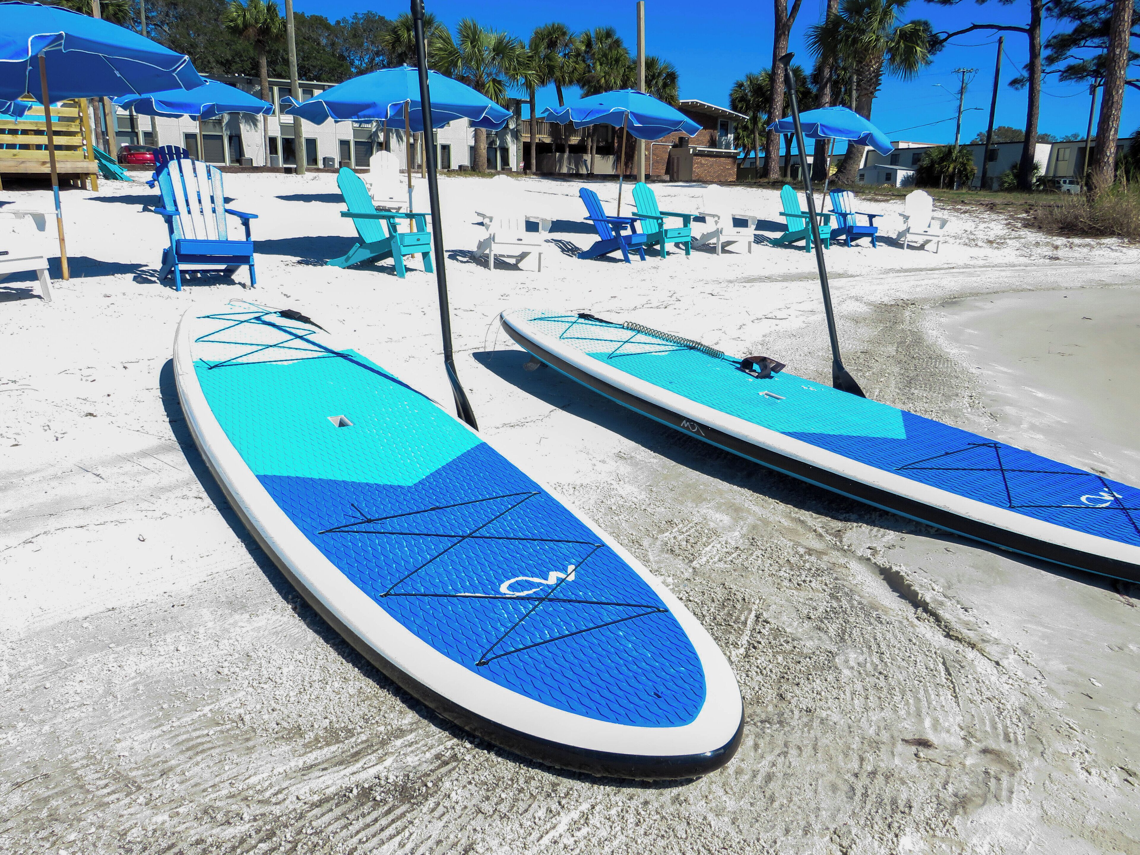 Dos tablas de paddle surf en una playa arenosa con sombrillas y sillas de playa azules.