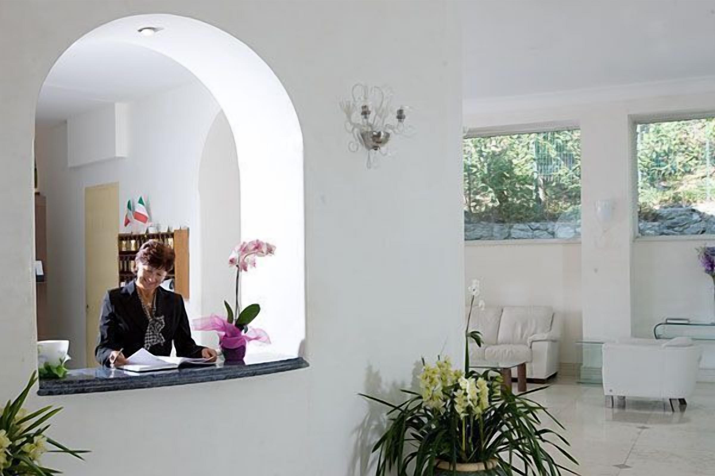 Et receptionsområde med receptionist, en receptionistskranke, en stor buegang, en stueplante og et siddeområde med hvid sofa og lænestole.