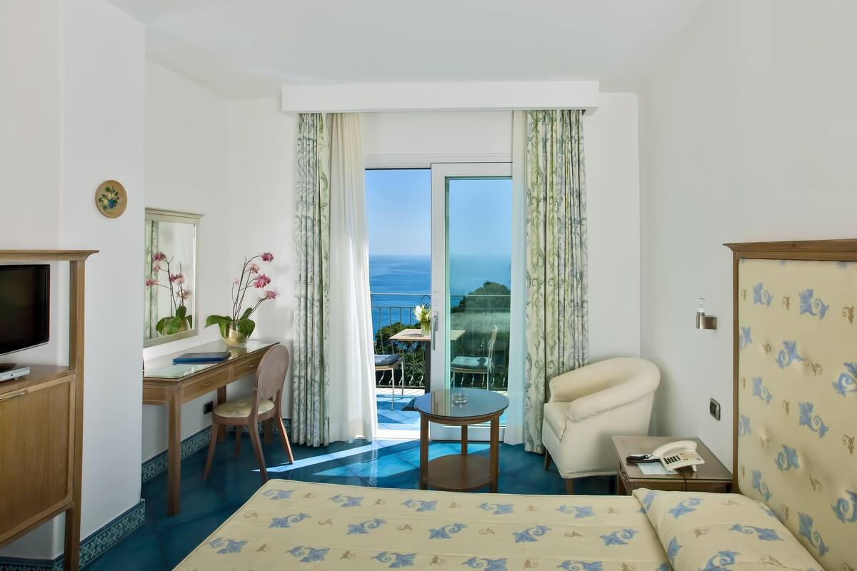 A hotel room with a bed, a desk, a chair, a small table, a TV, and a balcony with a view of the sea.