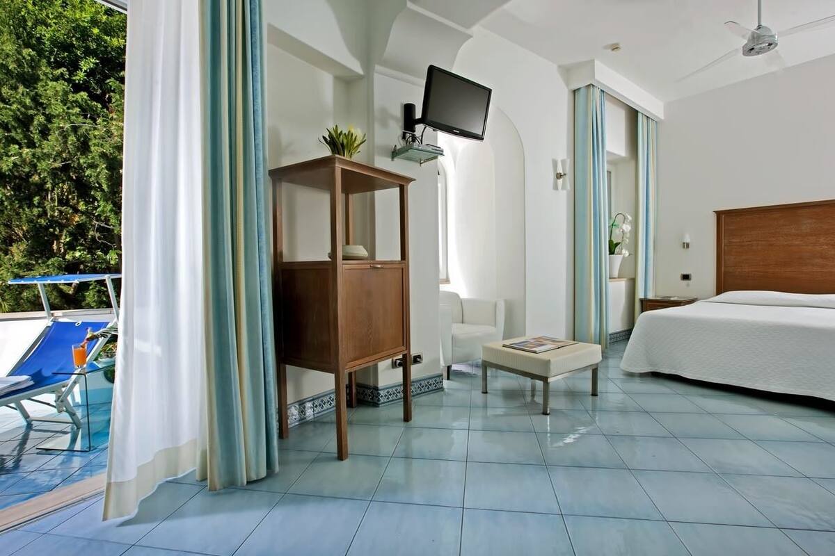 A hotel room with a bed, a small table, a TV, and a balcony with a chair.