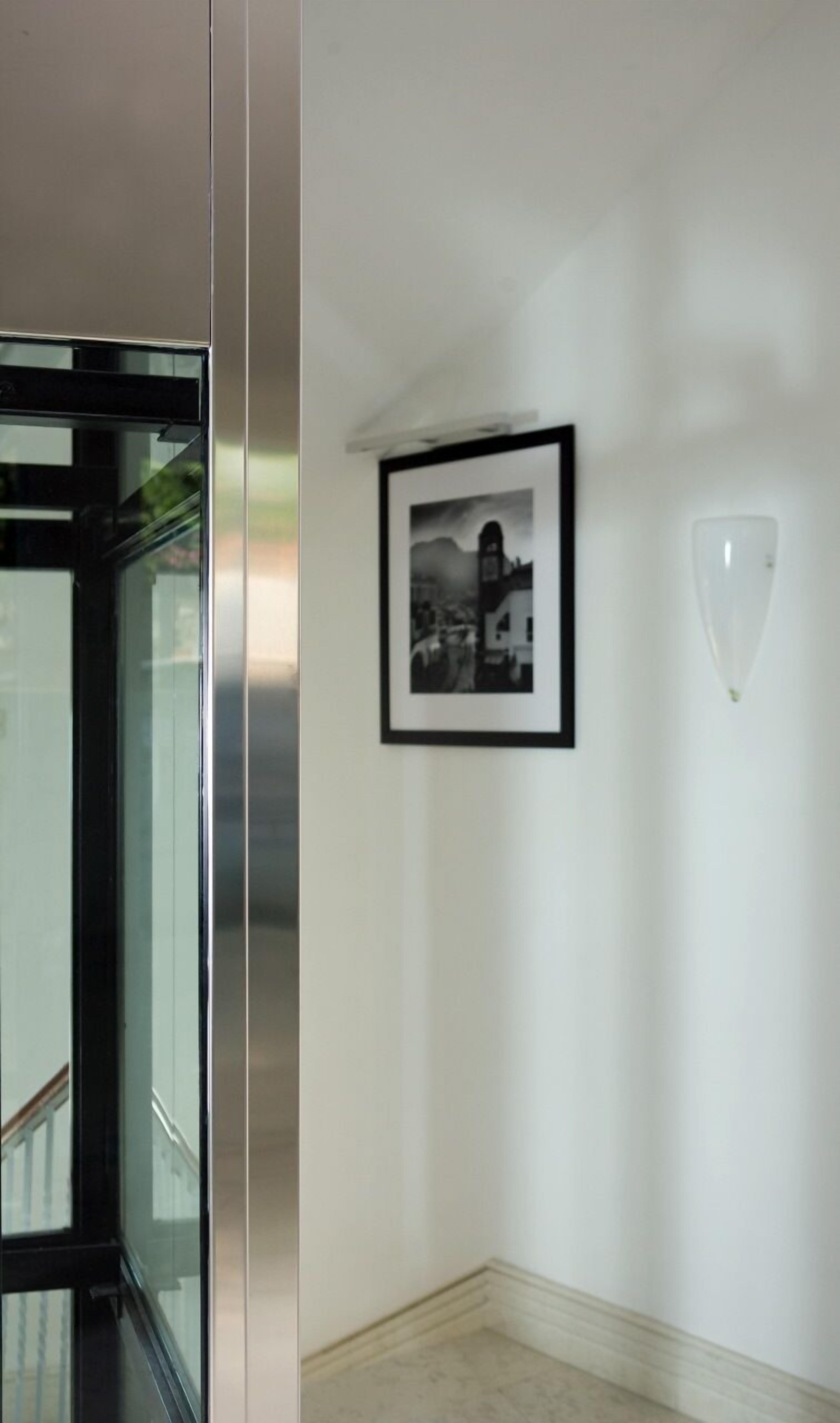Ein Flur mit einem in Schwarzweiß gerahmten Foto an der Wand, einer Leuchte und einer Glastür zu einem Aufzug.