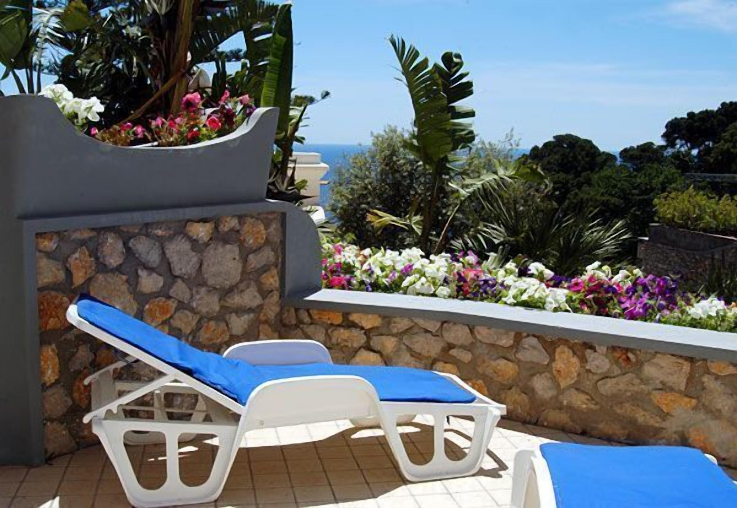 A sunlit outdoor lounge area with two white lounge chairs and a stone wall backdrop.
