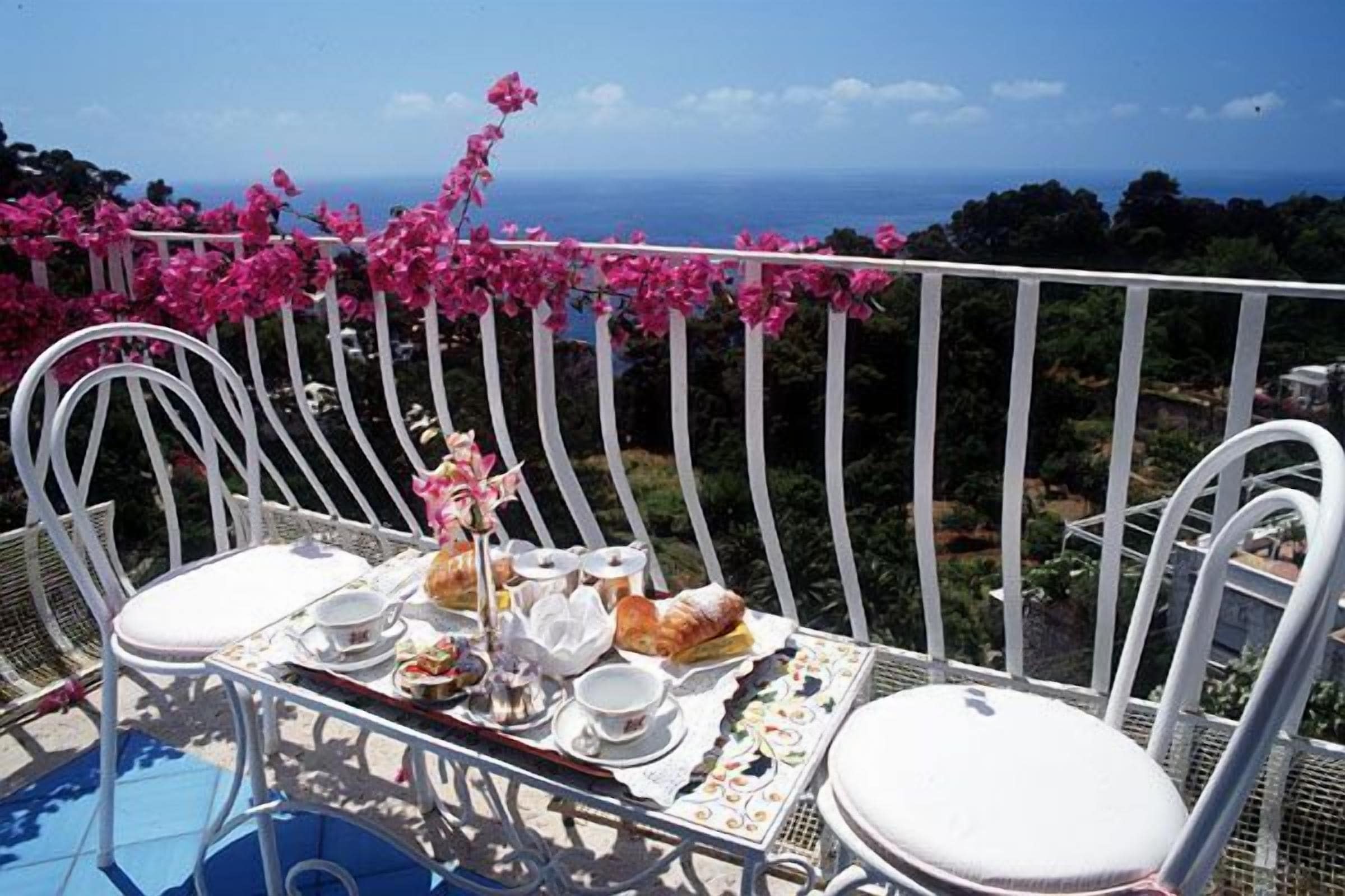 A balcony with a table set for tea, surrounded by white chairs, overlooking a scenic view with pink flowers and a distant landscape.