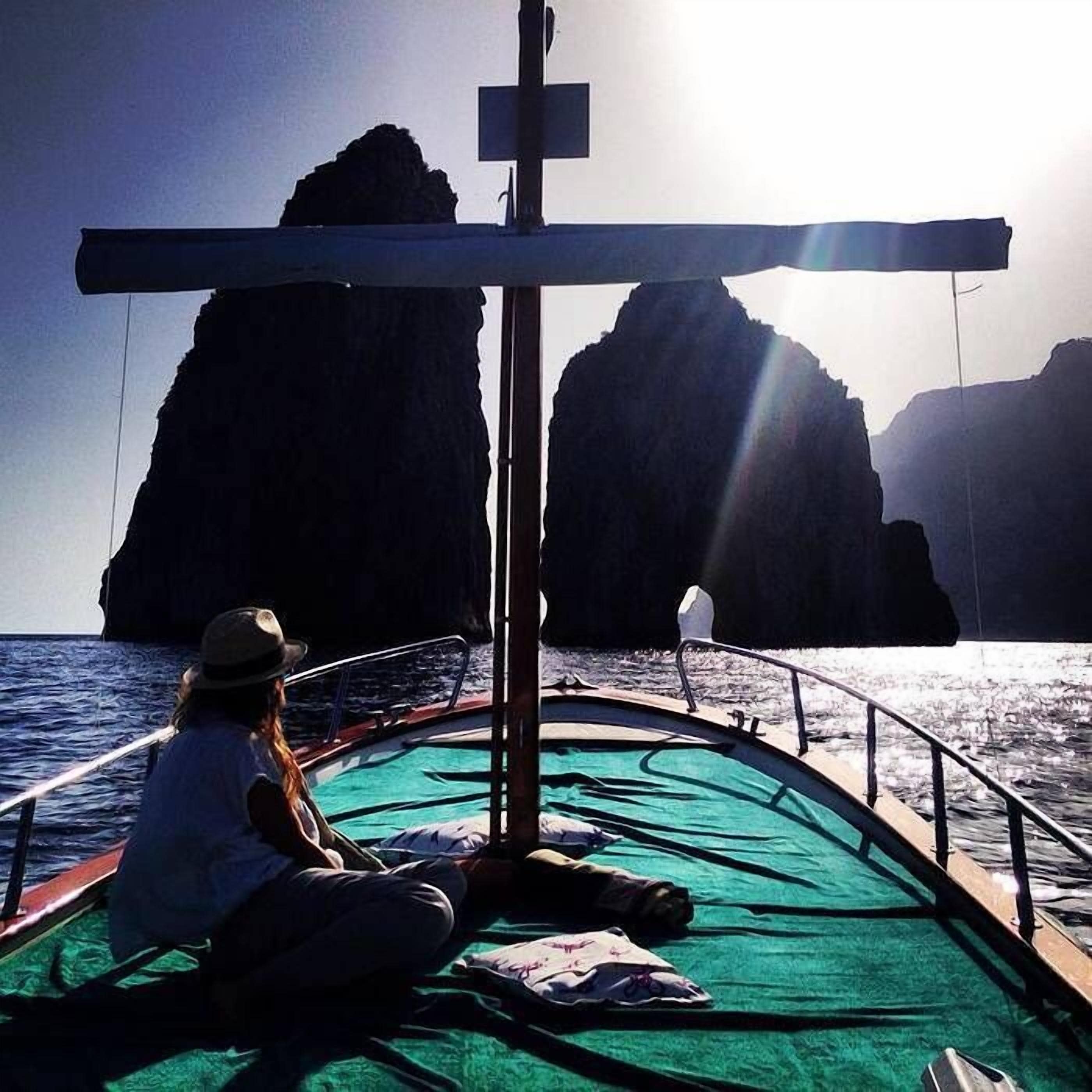 A boat with a person sitting on the deck, a cross structure, and large rock formations in the background.