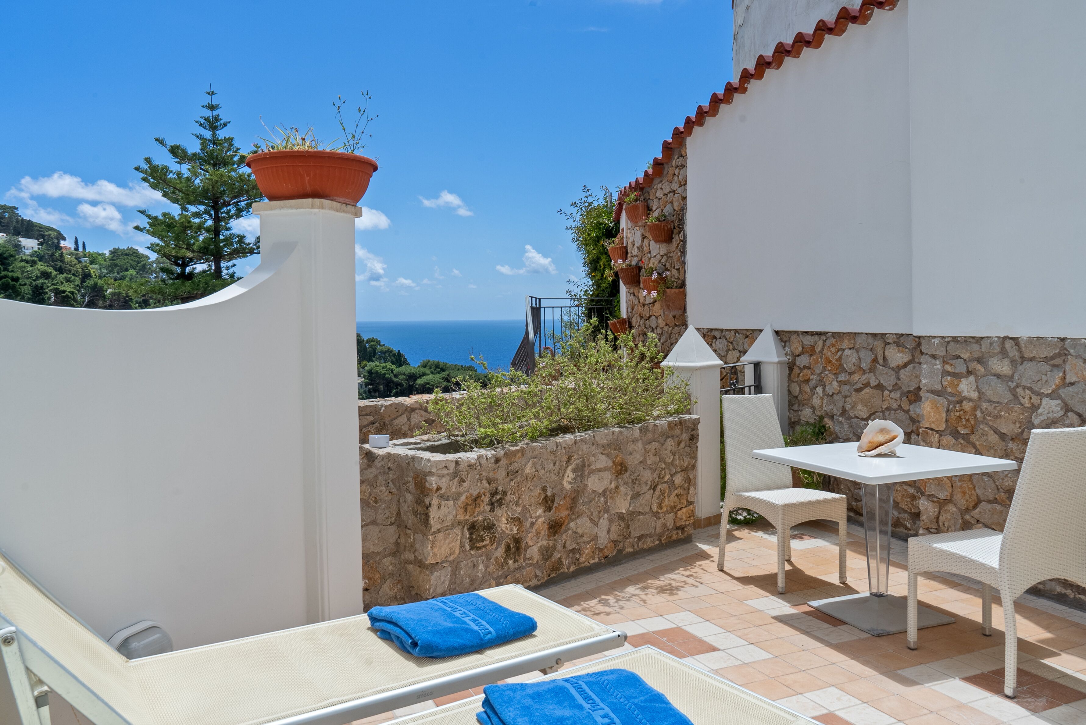 A terrace with a white table and chairs, a white lounge chair, and a view of the sea.