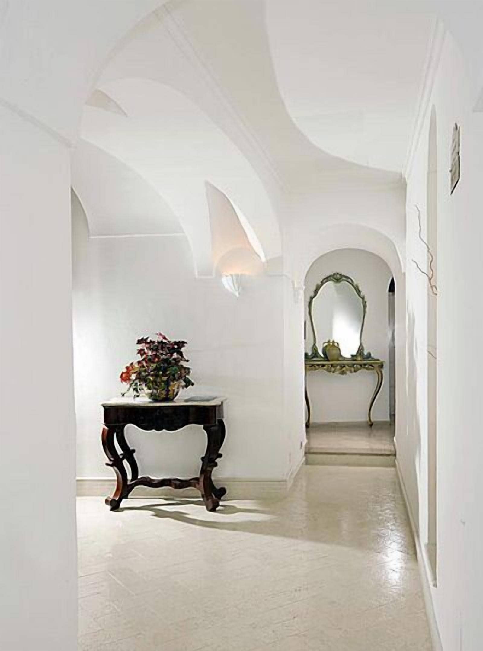 Un pasillo con paredes blancas y arcos, una pequeña mesa con un jarrón de flores y un espejo en la pared.