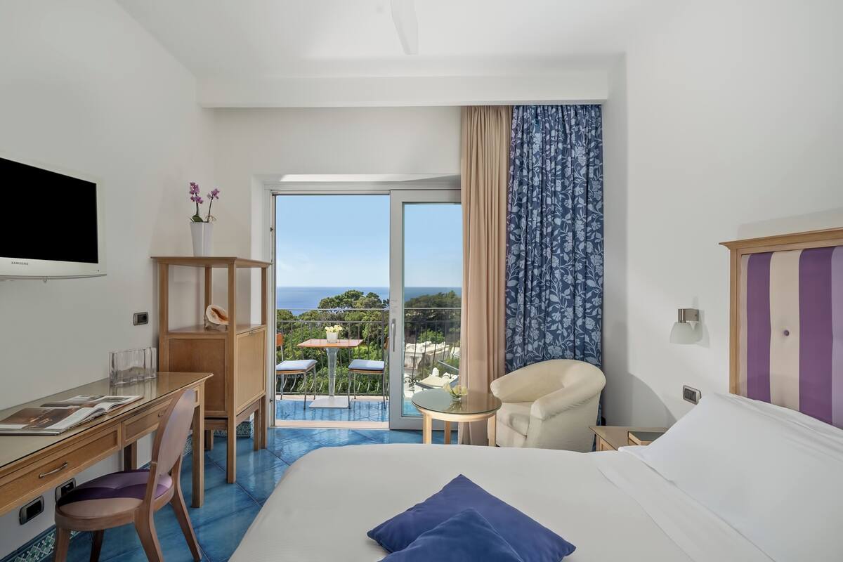 A hotel room with a bed, desk, chair, and a balcony with a view.