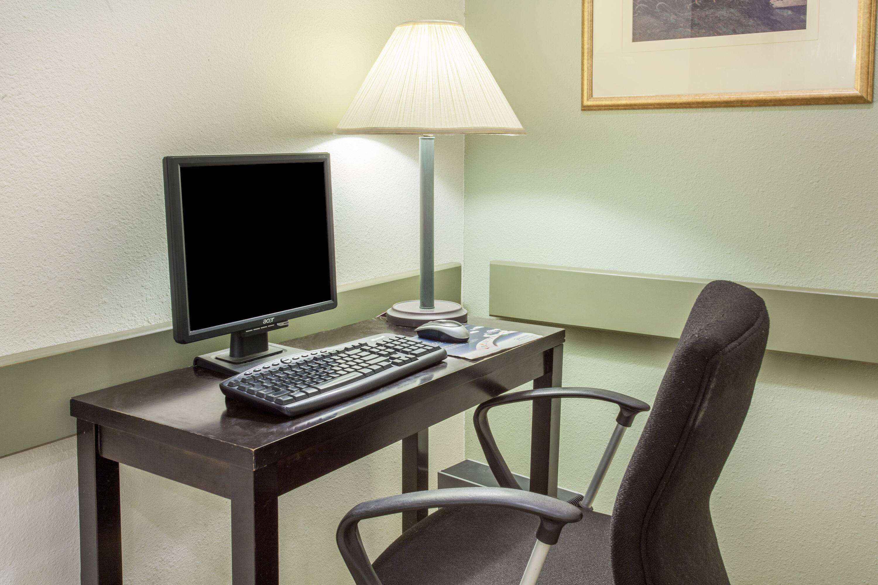 A hotel room with a desk, a computer monitor, keyboard, and a lamp.