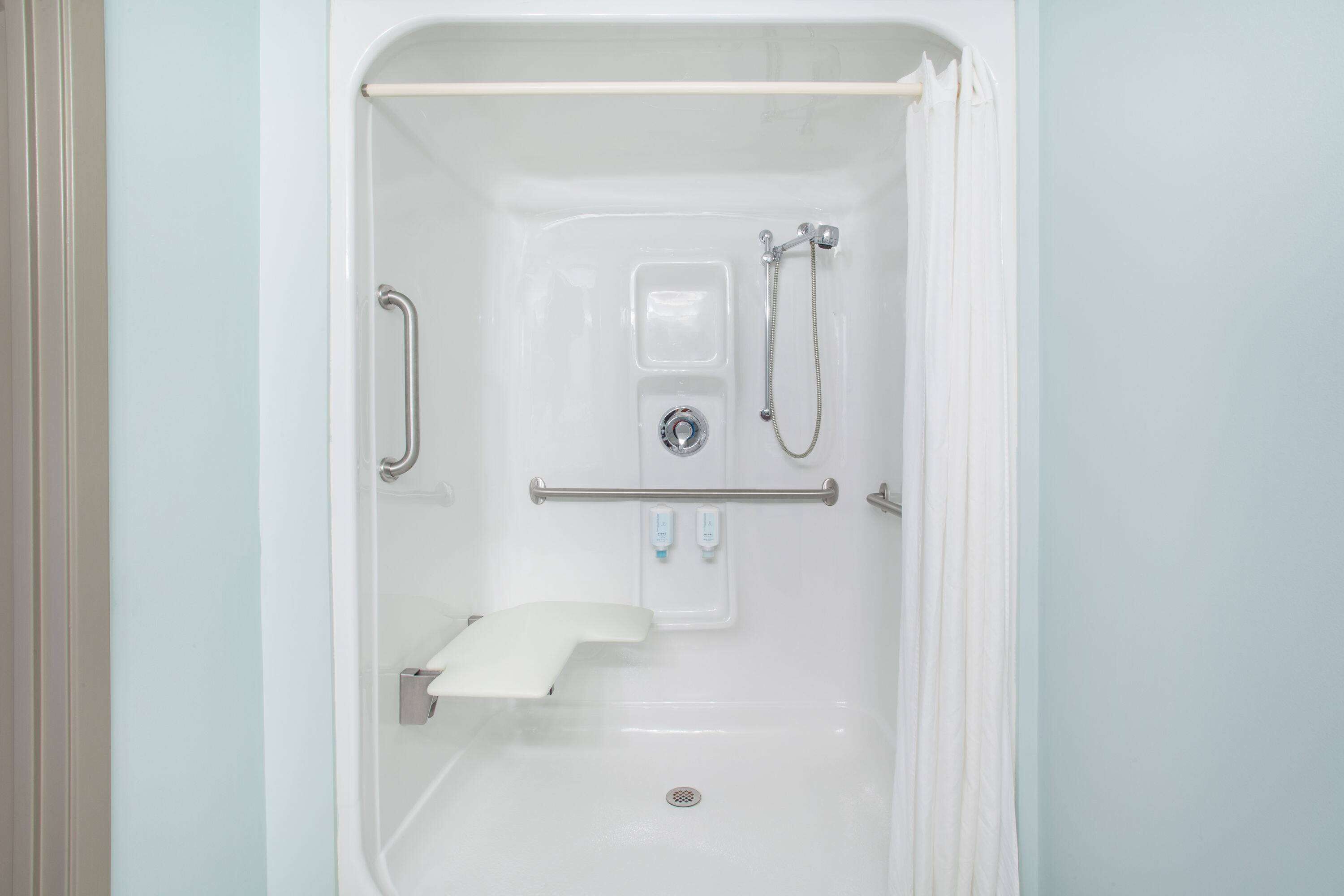 A white, accessible shower stall with a grab bar, handheld showerhead, and a handheld faucet.
