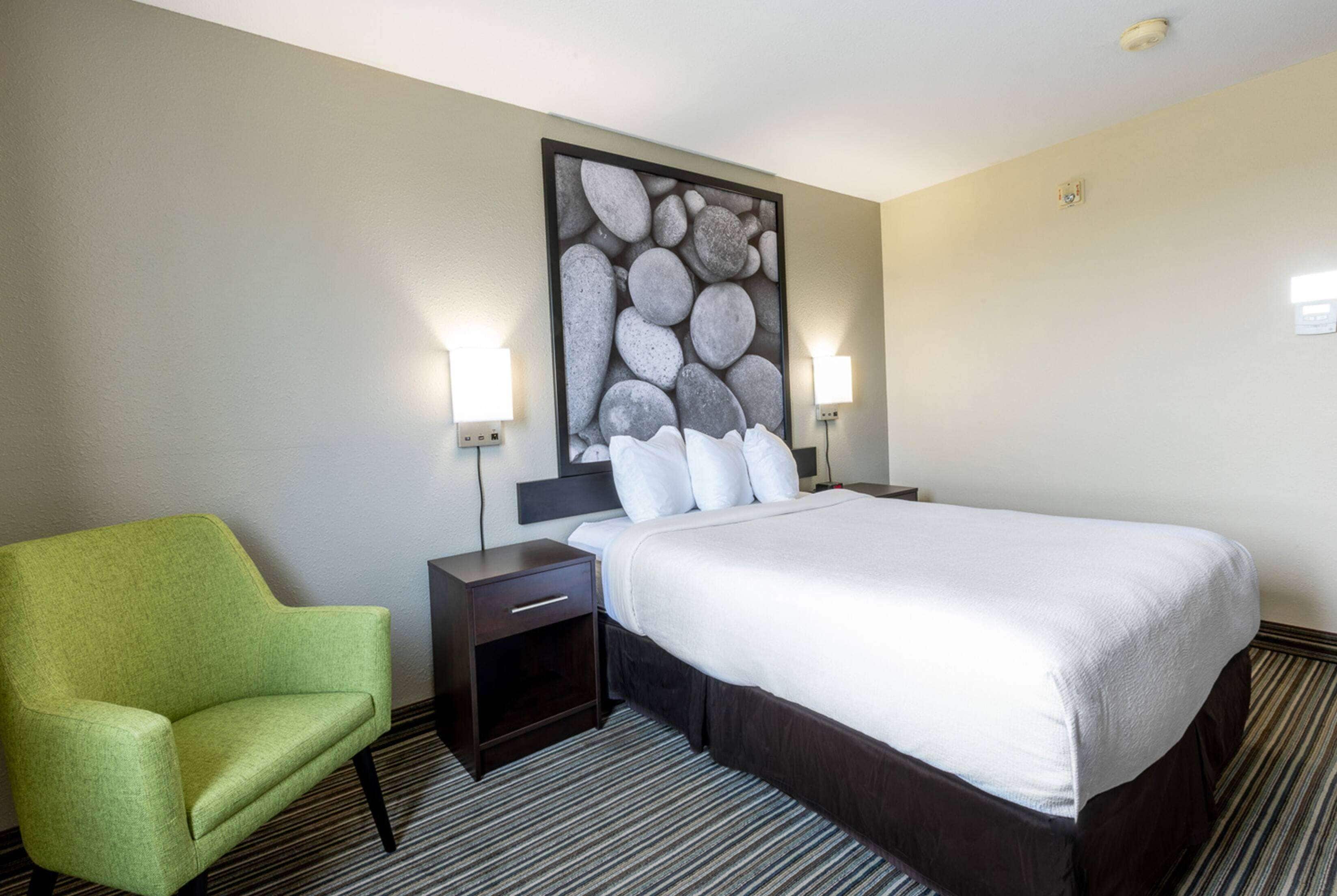 A hotel room with a large bed, a green armchair, two bedside tables with lamps, and a wall-mounted artwork.