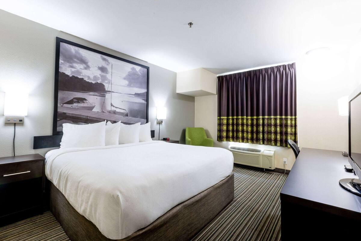 Habitación de hotel con una cama grande, un escritorio, una silla, un ventanal con cortinas, aire acondicionado de pared y un cuadro en la pared.