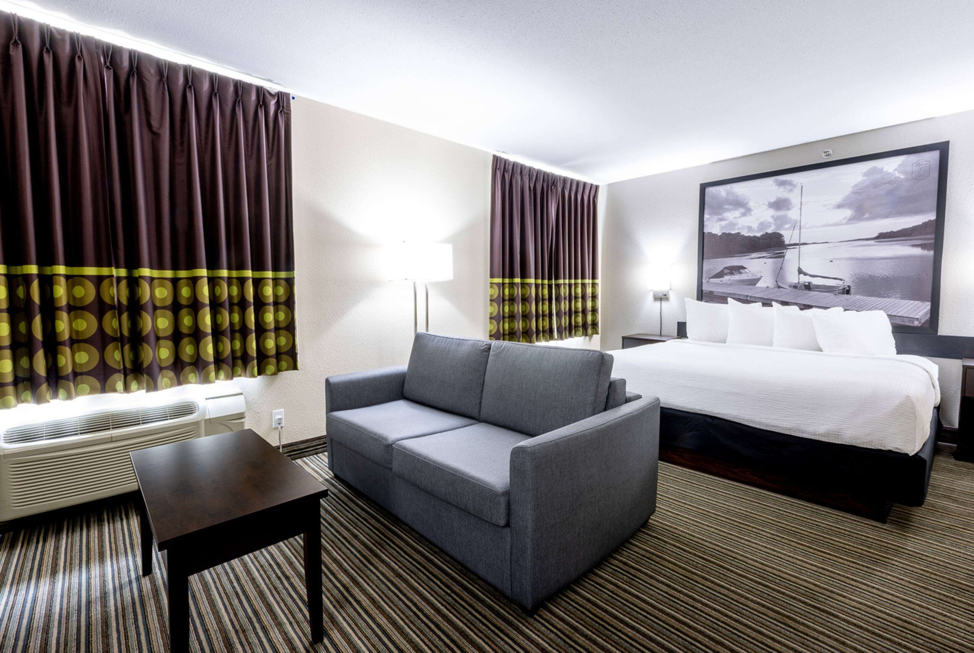 Habitación de hotel con sofá gris, una mesita pequeña, cama con ropa de cama blanca, ventana grande con cortinas y un televisor de pared que muestra una imagen panorámica.