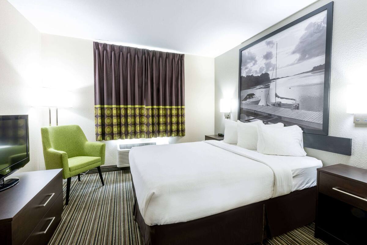 Habitación de hotel con cama, una silla, escritorio con televisor, ventana con cortinas y un cuadro en la pared.