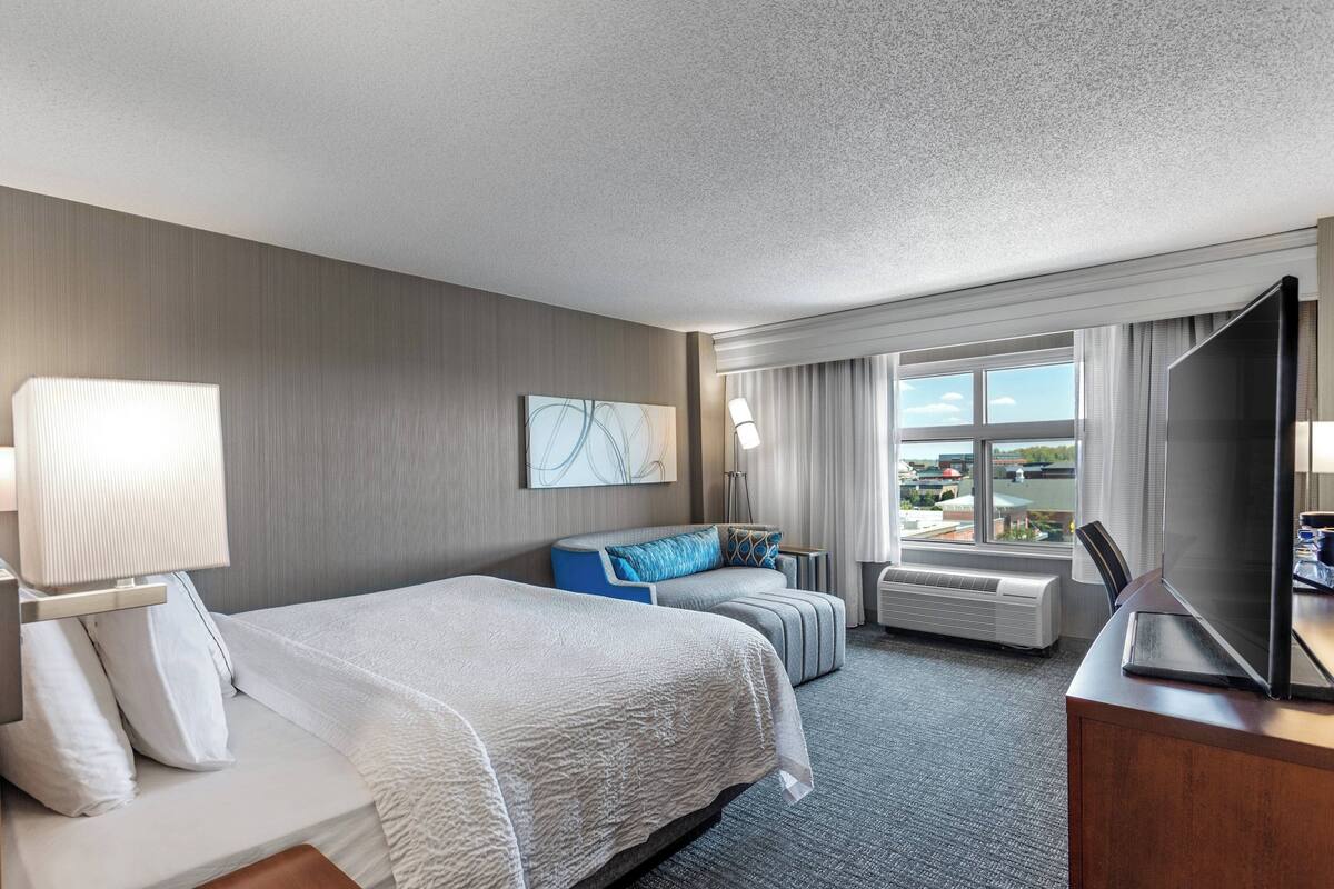 A hotel room with a large bed, a smaller sofa, a desk with a TV, and a window with a view.
