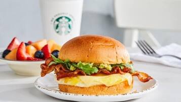Ein Frühstücksbrötchen mit Speck, Salat und Ei, eine Starbucks-Tasse und eine Schale Obst im Hintergrund.