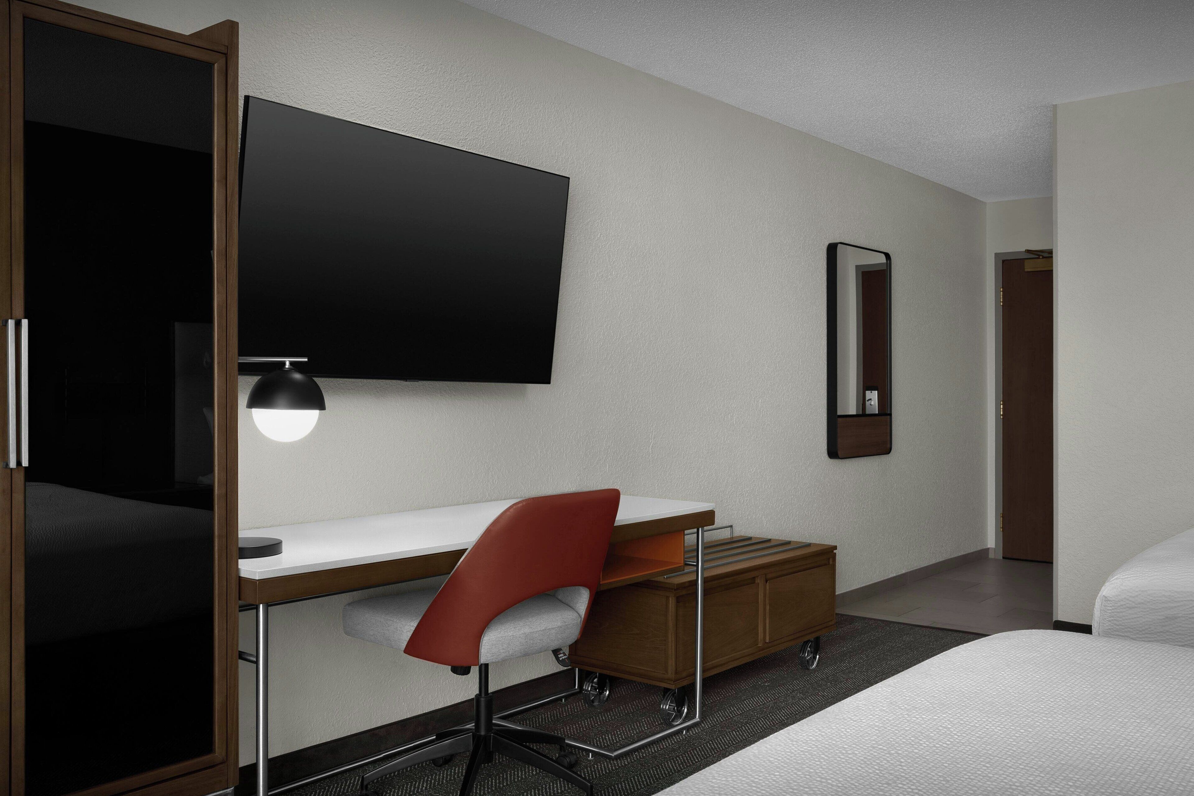 Ein Hotelzimmer mit einem Bett, einem Schreibtisch mit Stuhl, einem Fernseher, einem Spiegel und einem Holzschrank.