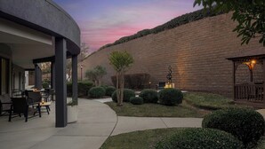 Un patio al aire libre con mobiliario y una pérgola, rodeado de setos cuidados y un muro de ladrillo.