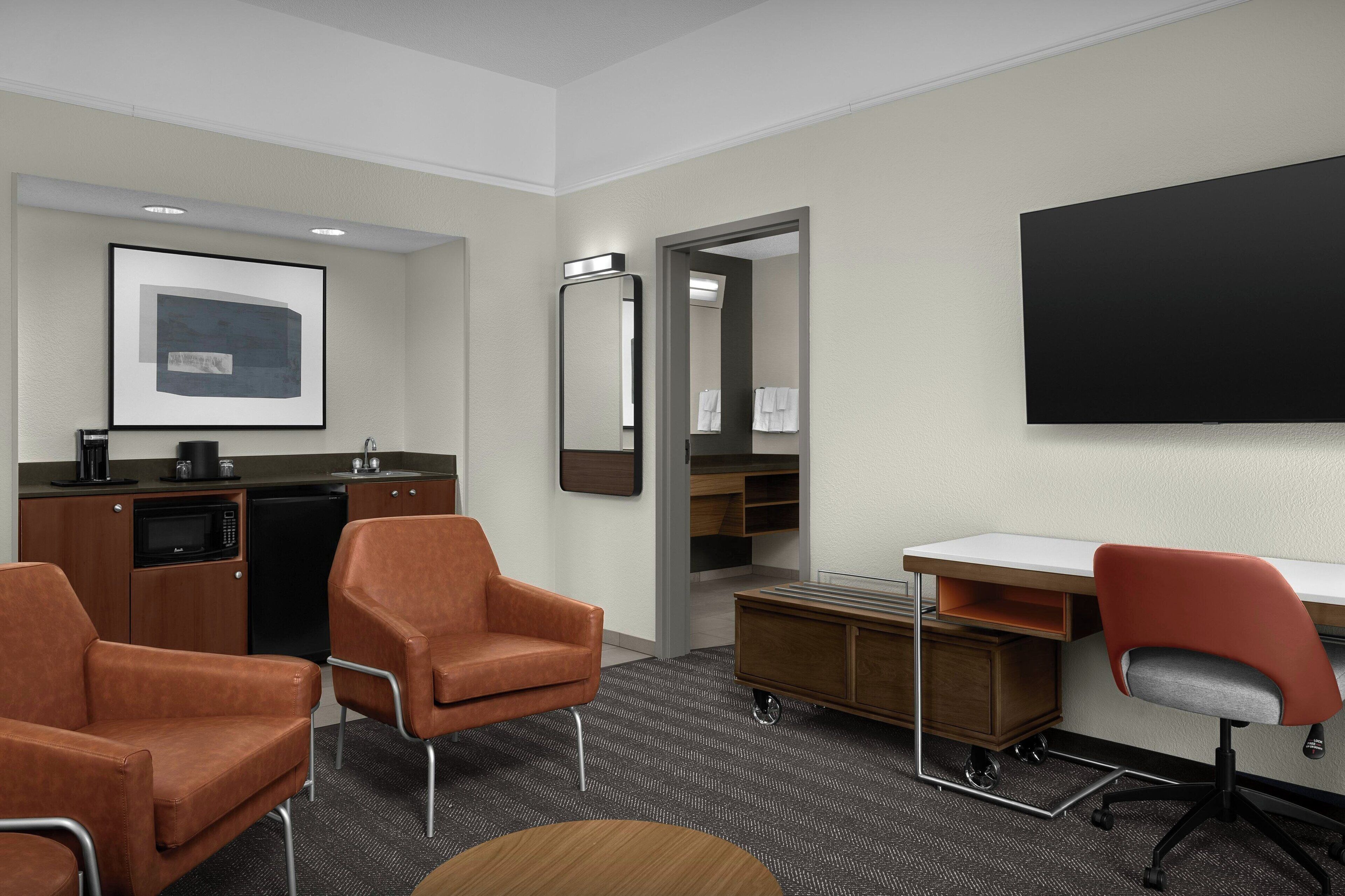 Een hotelkamer met een kitchenette, twee oranje stoelen, een bureau met stoel, een grote spiegel en een flatscreen televisie.