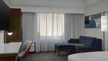 Une chambre d’hôtel comprenant un lit, un bureau, une chaise, un canapé, une petite table, une lampe, un tableau et une fenêtre avec des rideaux.
