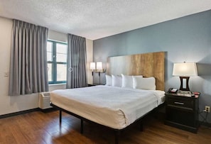 A hotel room with a large bed, a nightstand with a lamp, a window with curtains, and a wall-mounted headboard.