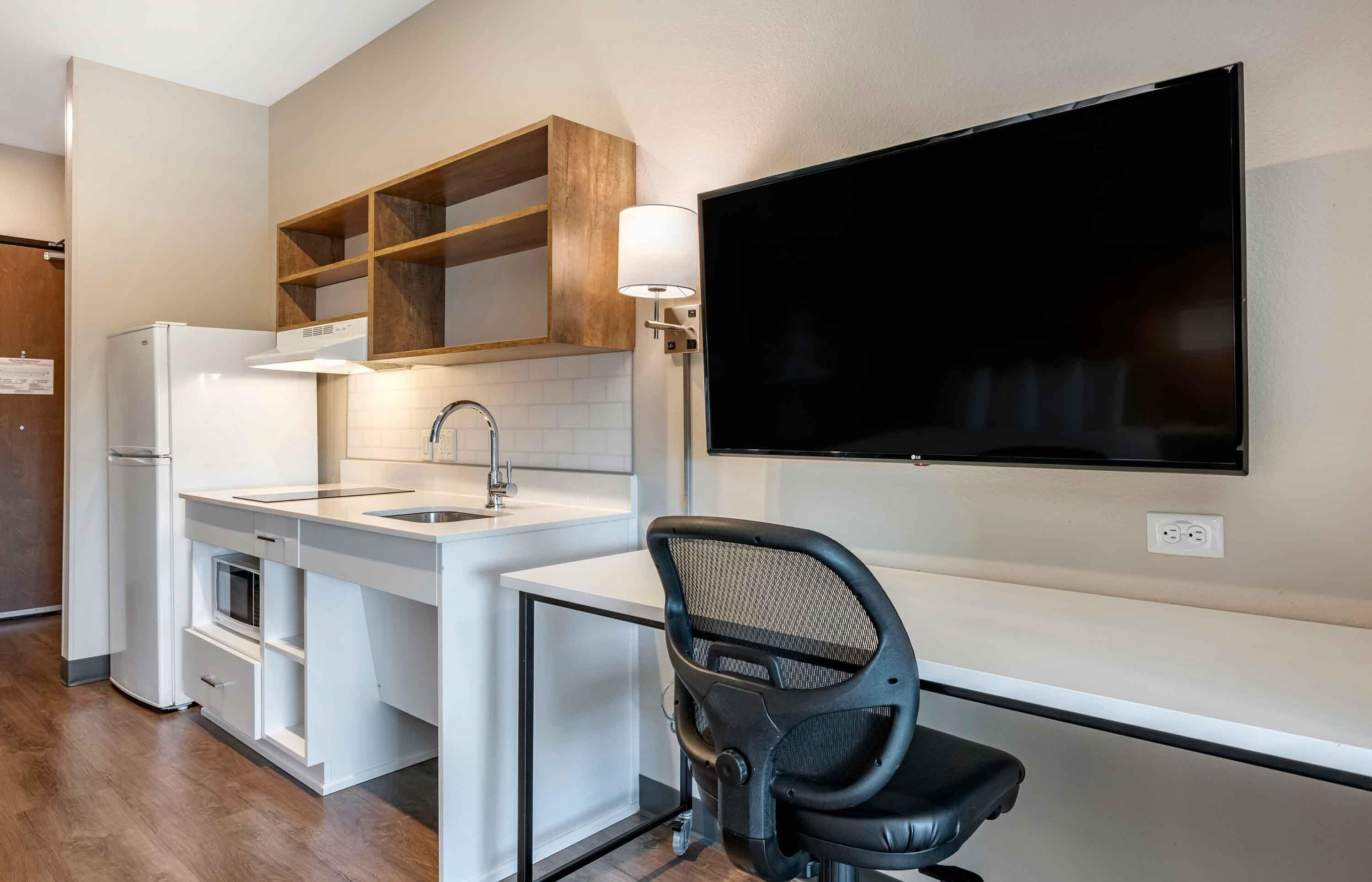 Een kitchenette met een koelkast, magnetron en spoelbak; een zwarte bureaustoel; en een flatscreen televisie aan de muur.