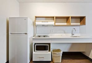 A compact kitchen with a refrigerator, microwave, and a sink with wooden shelves above.