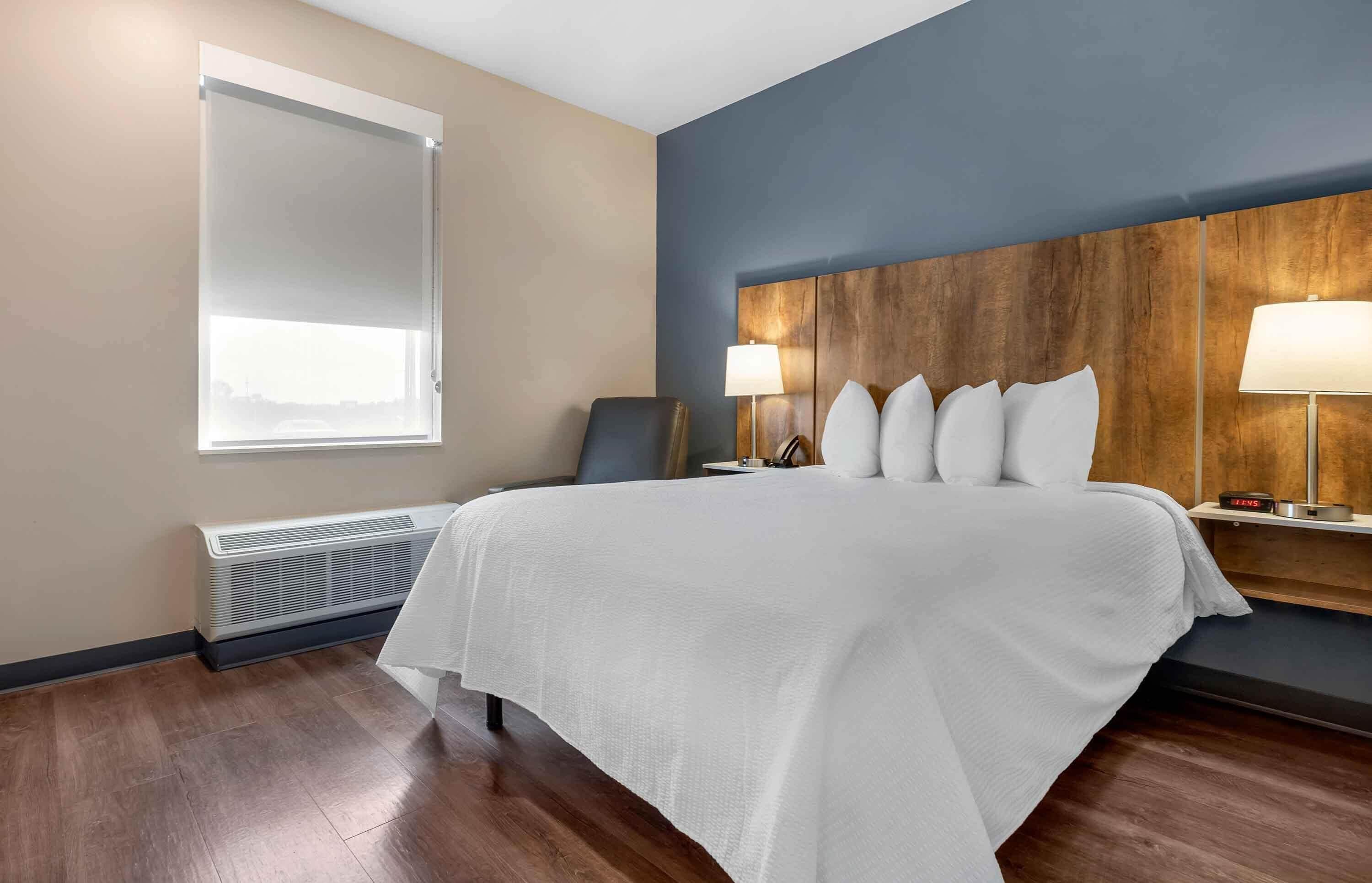 A hotel room with a large bed, a wooden headboard, two bedside lamps, a window with blinds, and a wall-mounted heater.